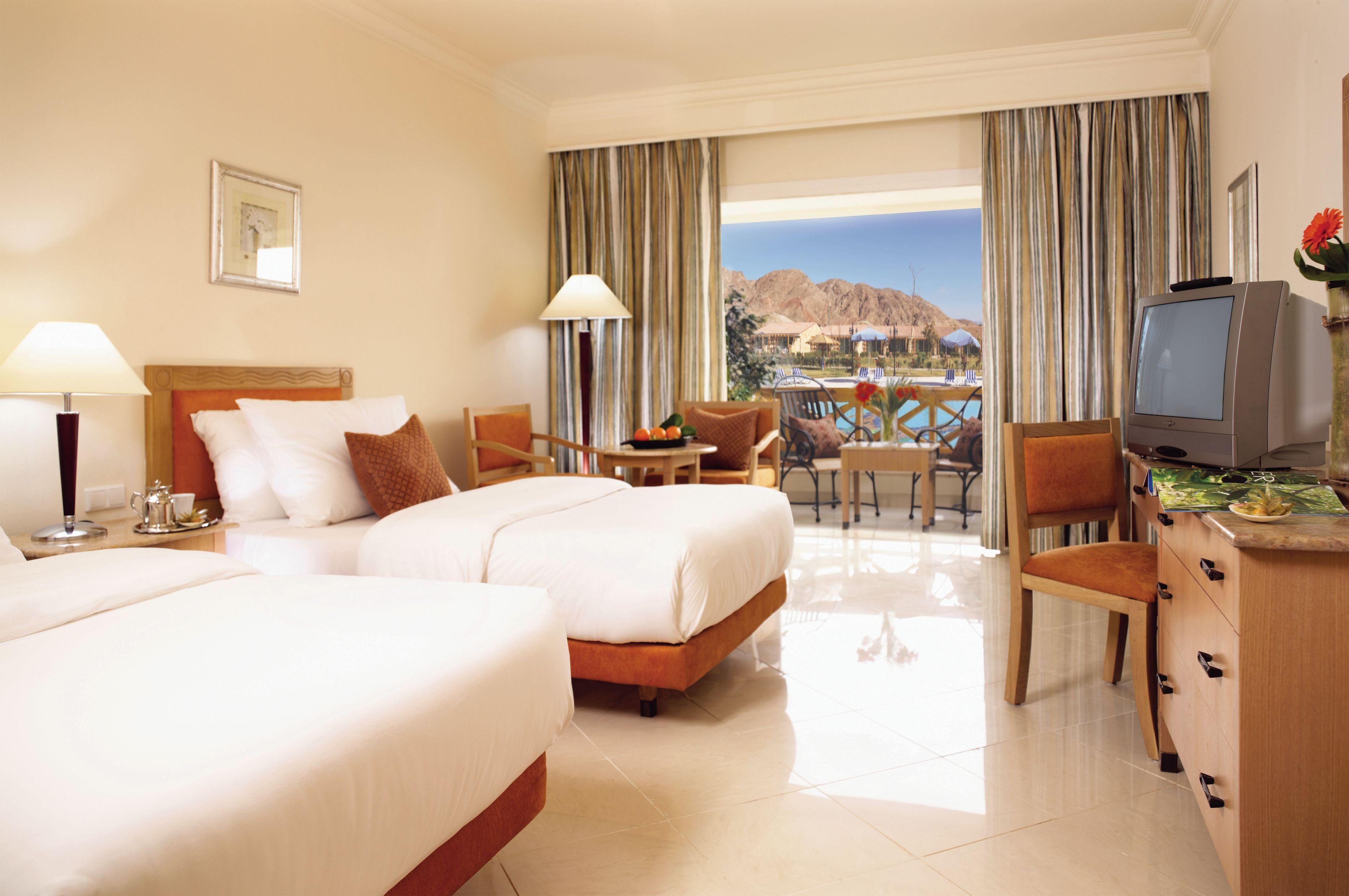 Movenpick Taba Resort & Spa Pokoj fotografie The photo shows a hotel room with two twin beds placed side by side. The bedding is light in color, and there are decorative pillows on each bed. A wooden bedside table with a lamp is positioned next to one of the beds. 

In the background, there are