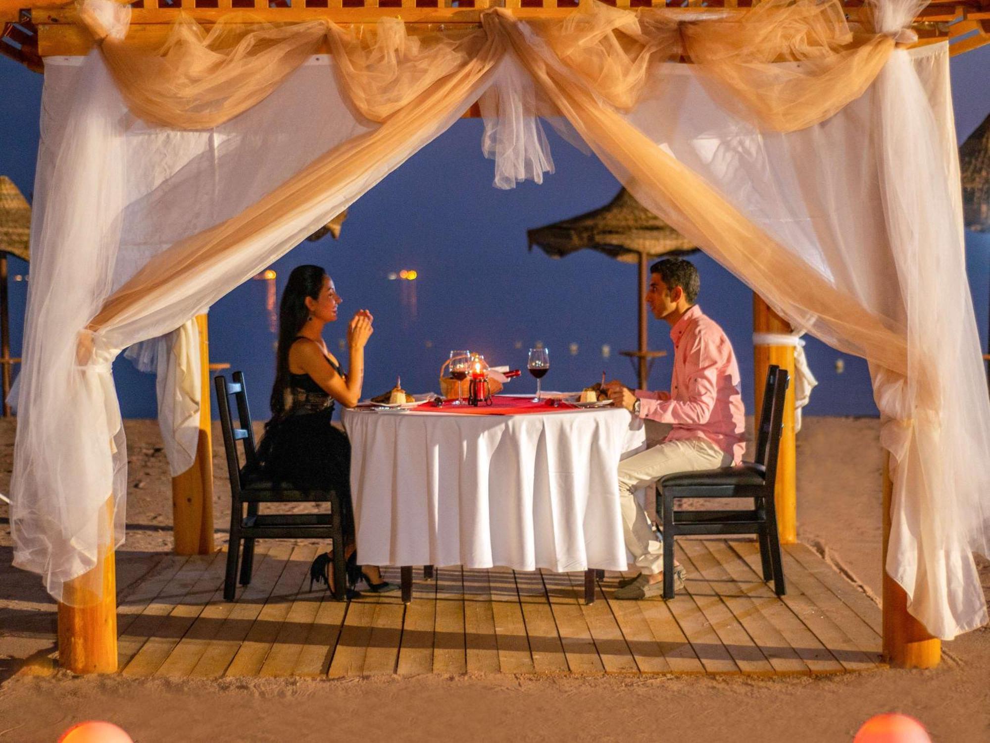 Movenpick Taba Resort & Spa Exteriér fotografie The photo depicts a romantic dining setup on the beach. A table is elegantly set with a white tablecloth and decor, including wine glasses and a centerpiece. There are two people seated at the table: one in a black dress and the other in a pink shirt